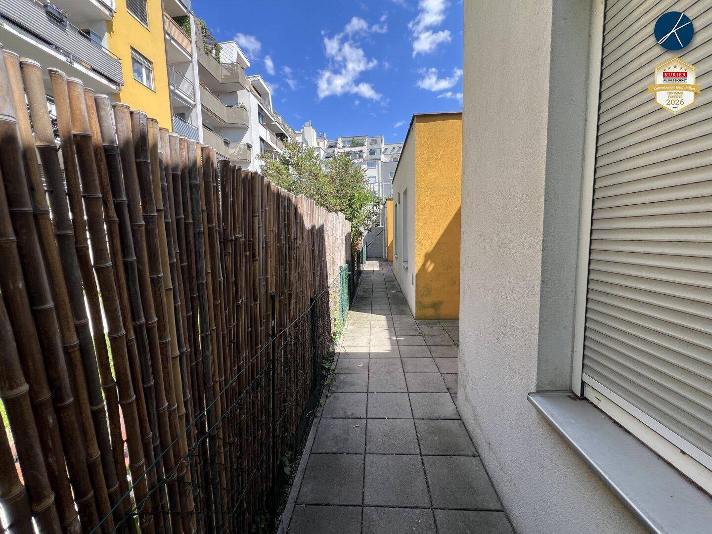 Schmaler gepflasterter Weg entlang einer Bambushecke zwischen Gebäuden unter blauem Himmel.