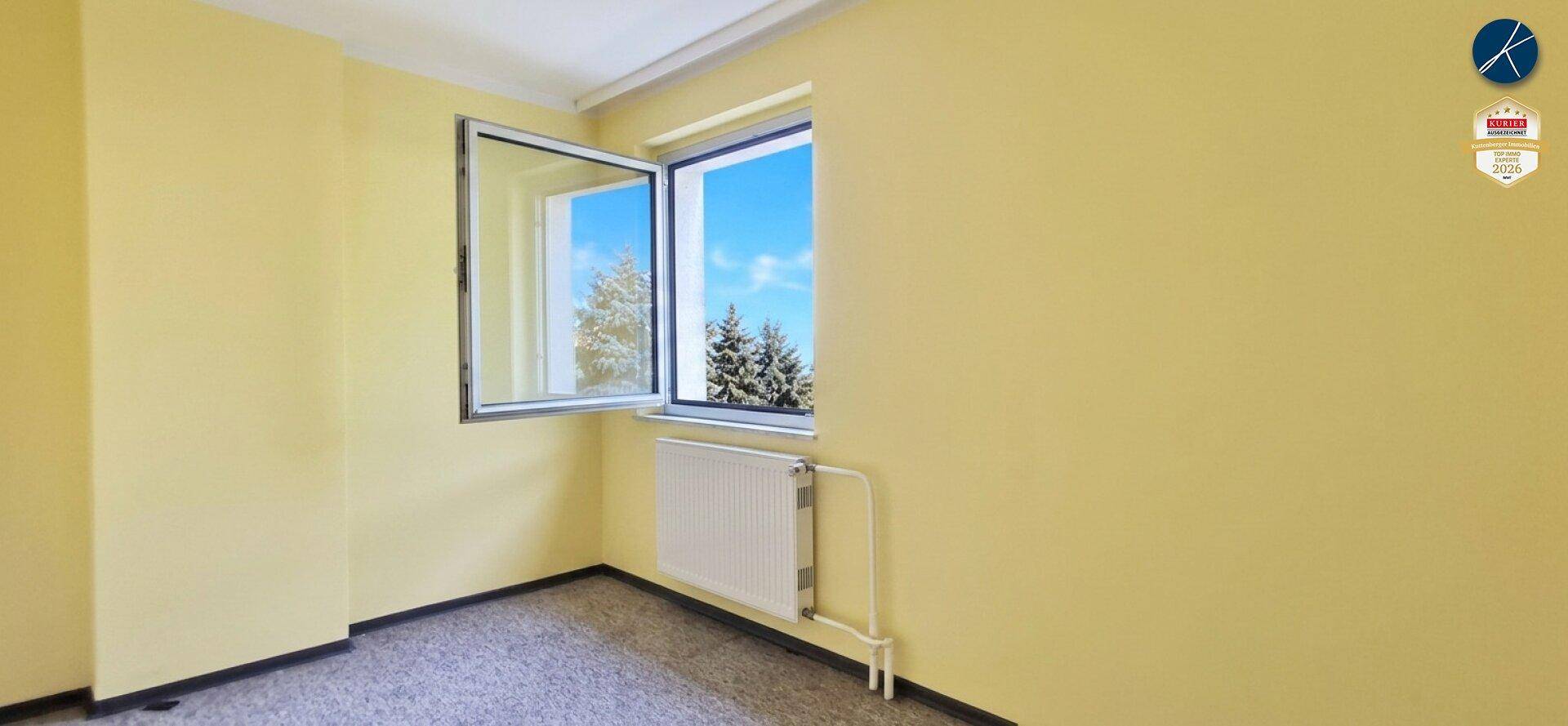 Helles Zimmer mit gelben Wänden, Teppichboden und einem Fenster mit Blick ins Grüne.