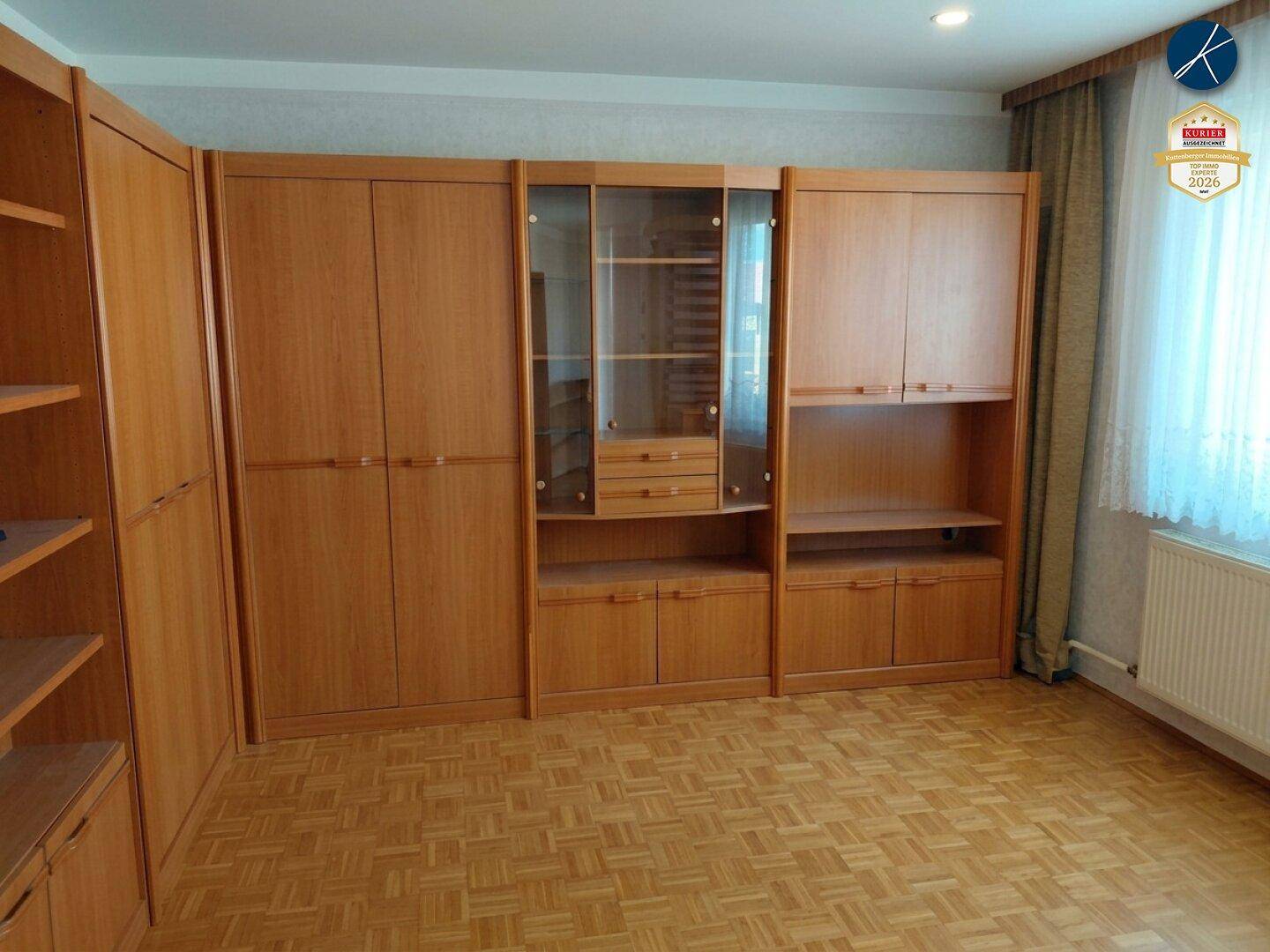 Wohnbereich mit umfangreichen Holzschränken, Parkettboden und Vorhängen am Fenster.