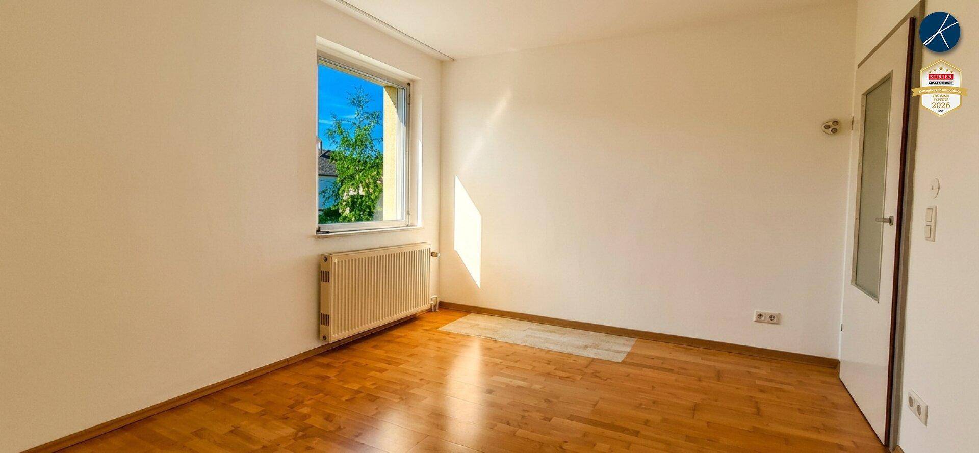Freundliches Zimmer mit Holzboden, weißen Wänden und viel Tageslicht durch das Fenster.