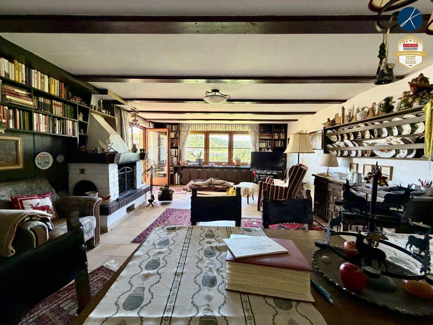 Gemütliches Wohnzimmer mit Kamin, Holzbalken und Bücherregalen, Blick ins Grüne.