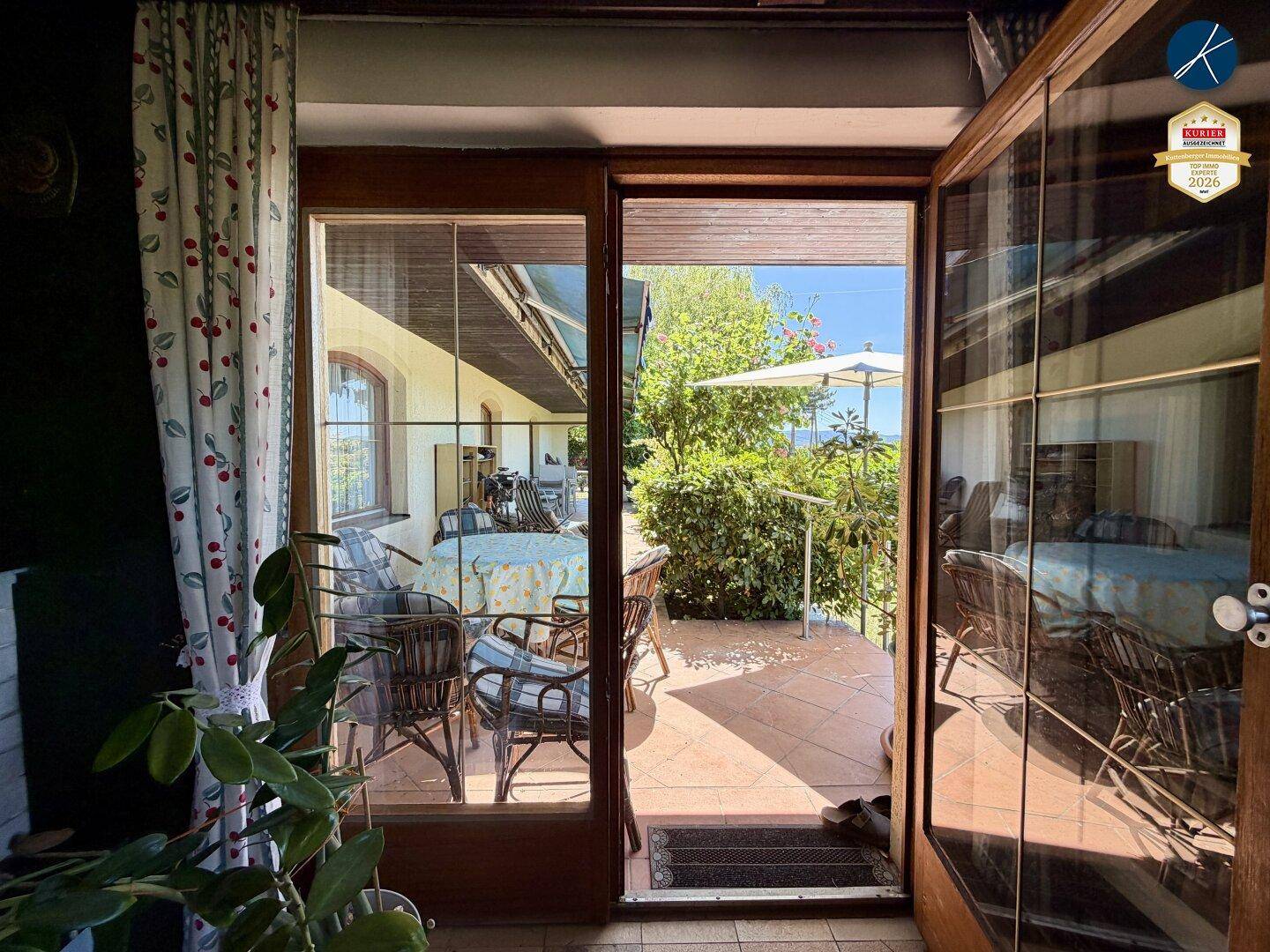 Blick aus dem Innenraum auf eine möblierte Terrasse mit Sonnenschirm und grüner Umgebung.