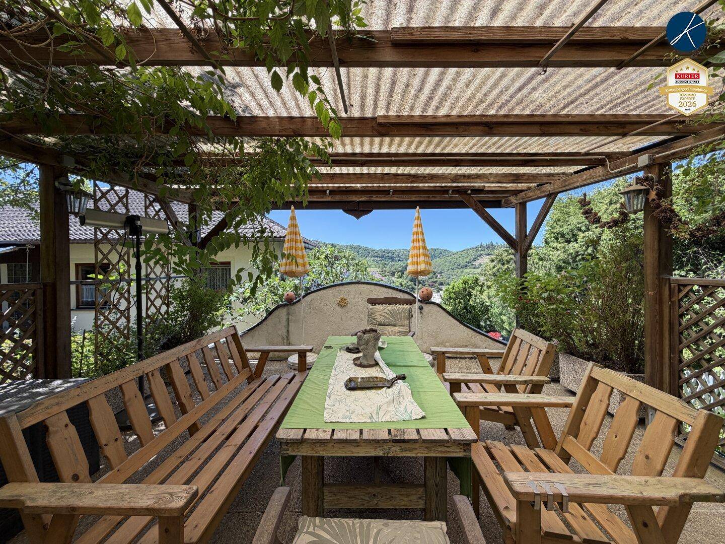 Überdachte Terrasse mit Holzbänken und Esstisch, umgeben von grüner Landschaft.