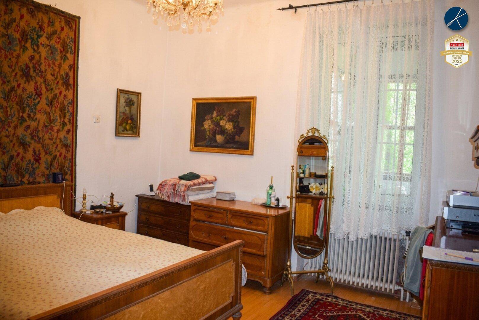 Schlafzimmer mit klassischem Holzbett, Kommoden und großem Fenster mit viel Tageslicht.