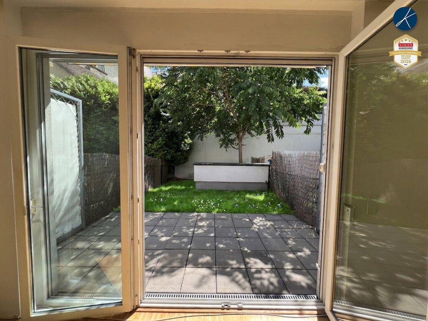 Blick vom Innenraum auf die gepflasterte Terrasse und den kleinen Gartenbereich mit Baum.