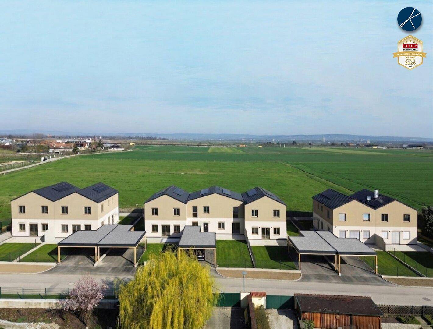 Luftaufnahme einer Reihe moderner Einfamilienhäuser mit Carports und Photovoltaikanlagen, umgeben von grünen Feldern.