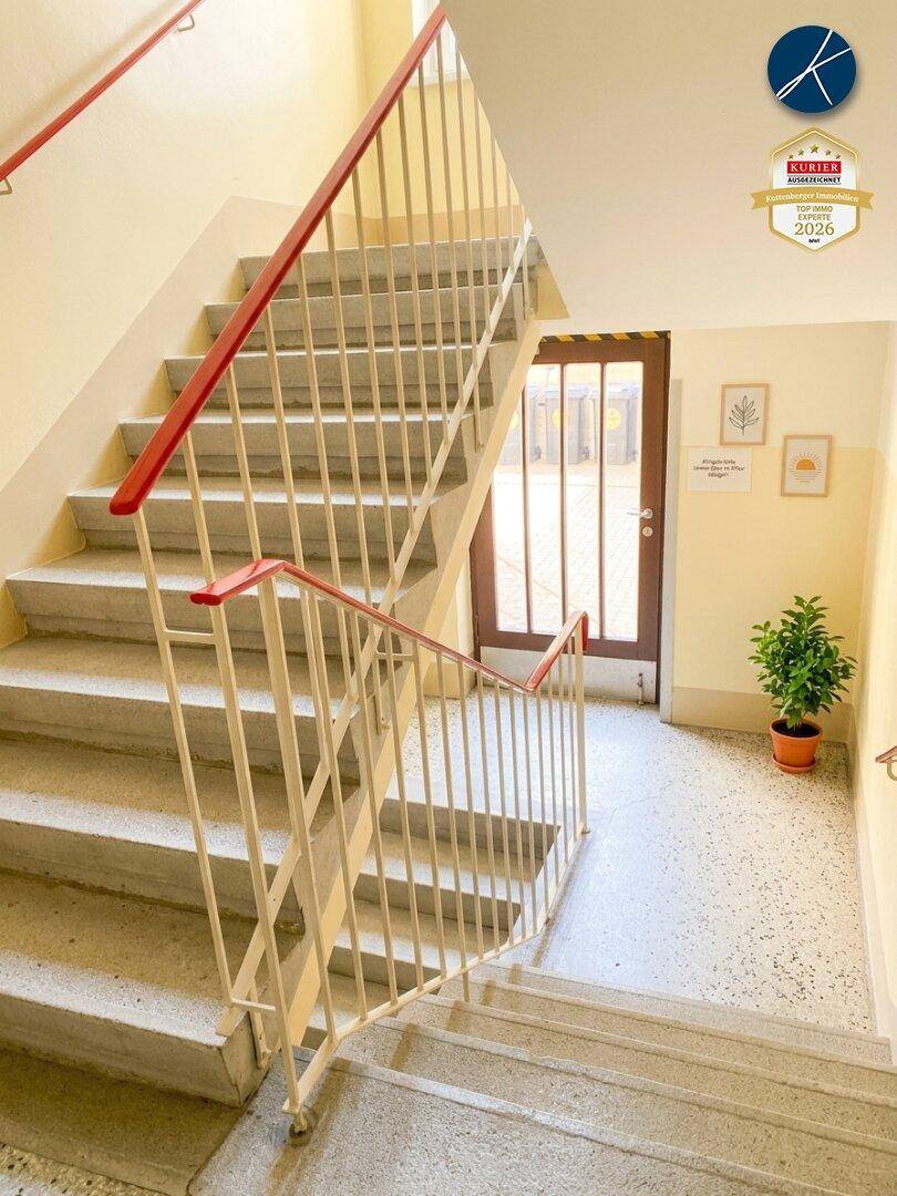 Helles Treppenhaus mit Terrazzoboden und roten Handläufen, das den Zugang zu den Wohnungen ermöglicht.