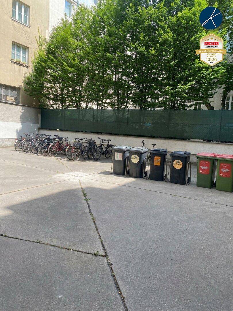 Hinterhofbereich mit Fahrradabstellplätzen und Mülltonnen, umgeben von Bäumen und einer Hecke.