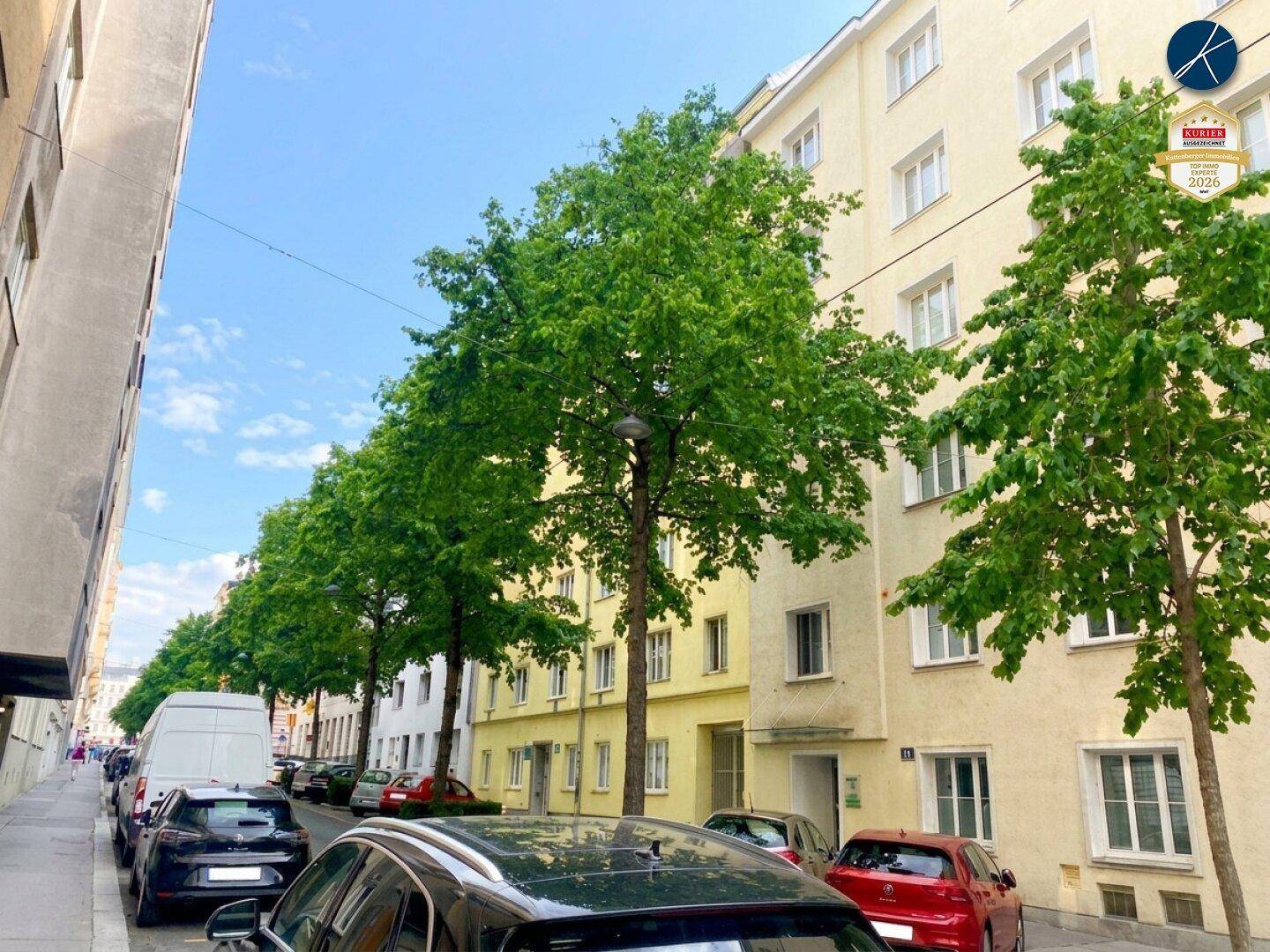 Ruhige Wohnstraße mit parkenden Autos und Bäumen, die Schatten spenden, vor einer Wohnhausfassade.