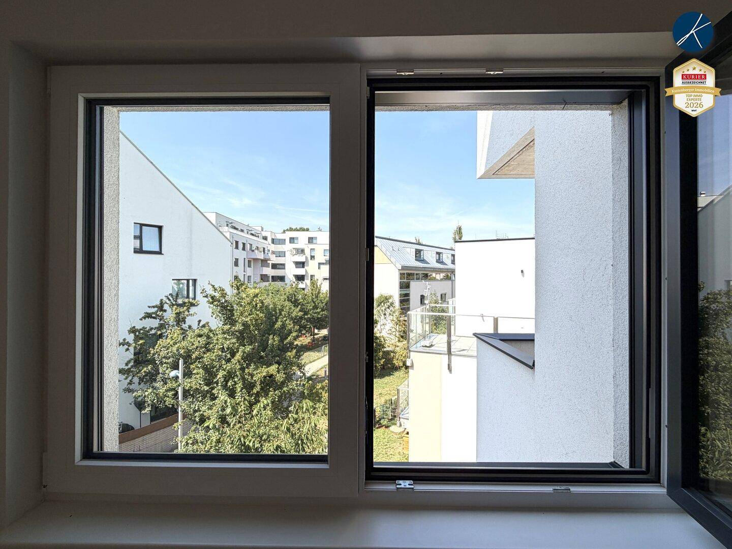 Blick aus einem Fenster auf die umliegenden Wohngebäude und grüne Bäume.