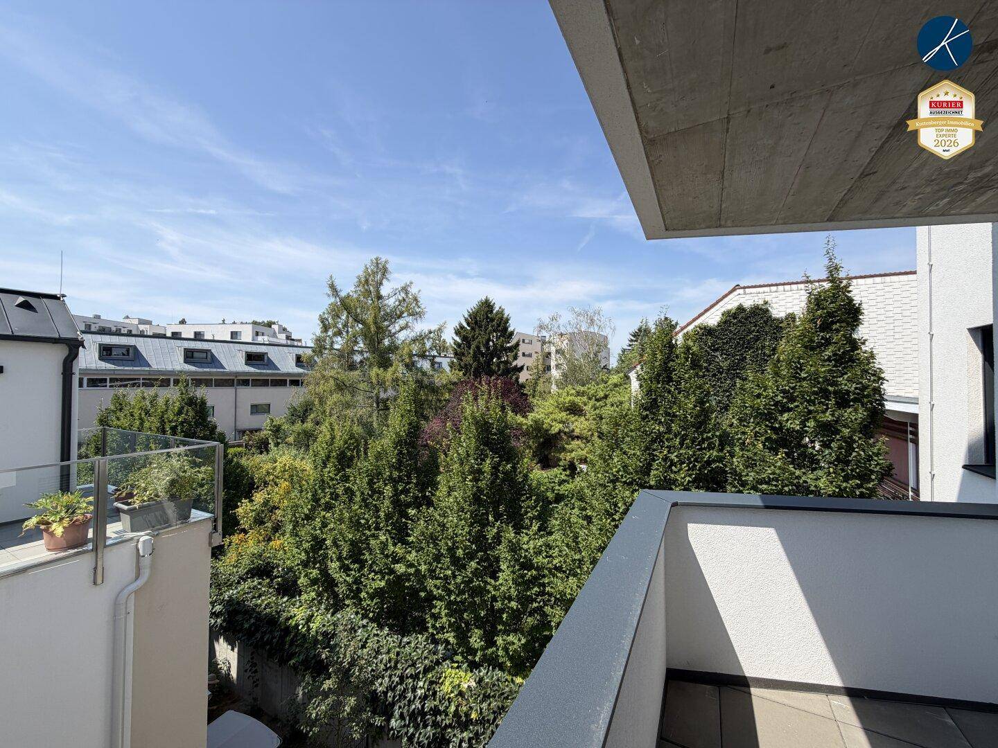 Balkon mit Blick auf grüne Bäume und umliegende Gebäude unter blauem Himmel.