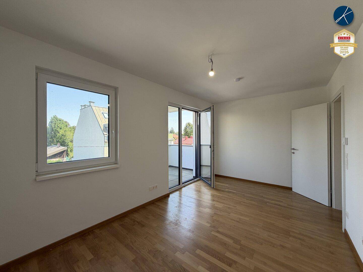 Helles Schlafzimmer mit Parkettboden, großem Fenster und Zugang zu einem Balkon.