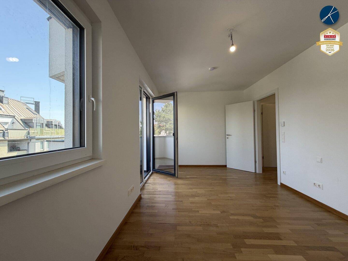 Lichtdurchflutetes Zimmer mit Holzboden, großem Fenster und offener Balkontür.