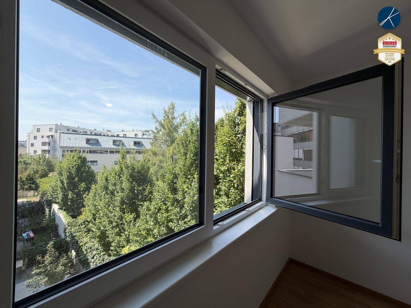 Fensterblick auf grüne Bäume und umliegende Gebäude unter blauem Himmel.