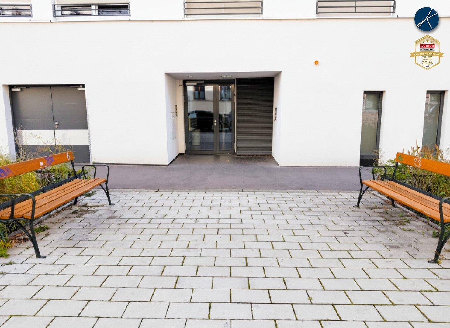 Eingangsbereich des Gebäudes mit gepflastertem Vorplatz und zwei Holzbänken.