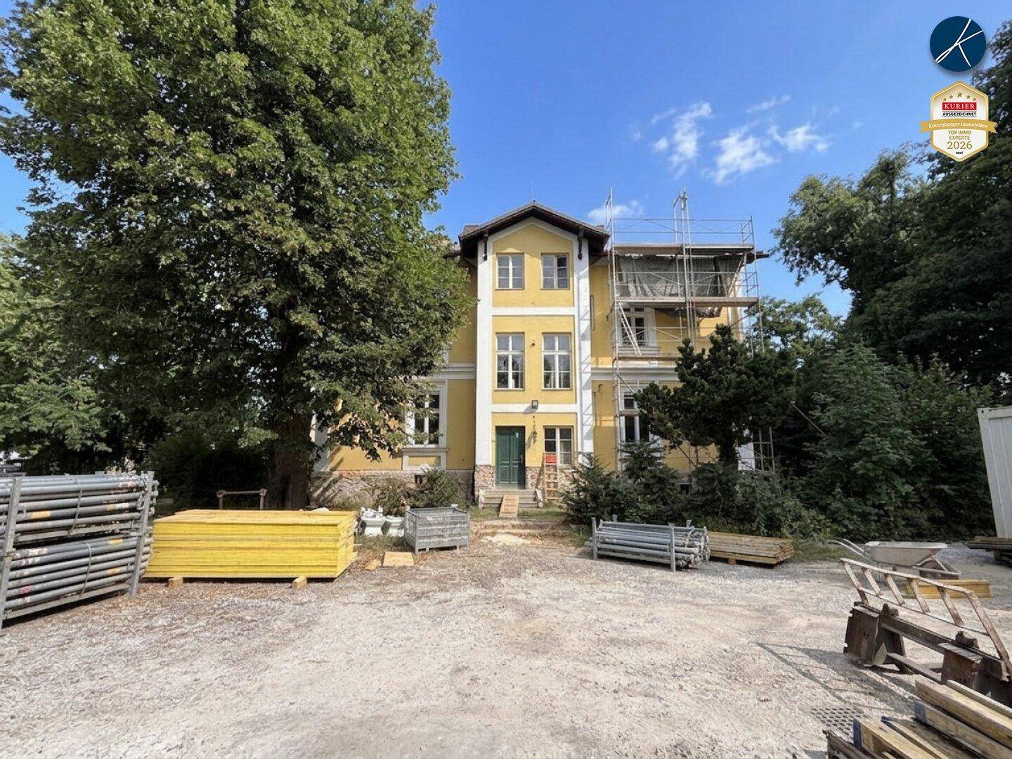 Gelbes Gebäude mit Baugerüst an der Seite, umgeben von Baustellenmaterialien und Bäumen.