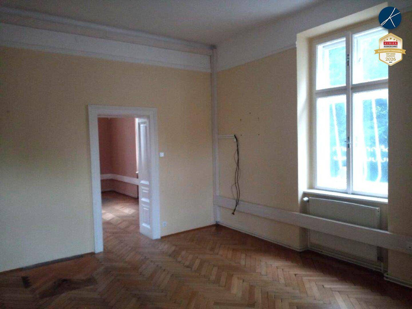 Leerer Raum mit Fischgrätparkett und großen Fenstern, die viel Tageslicht hereinlassen.