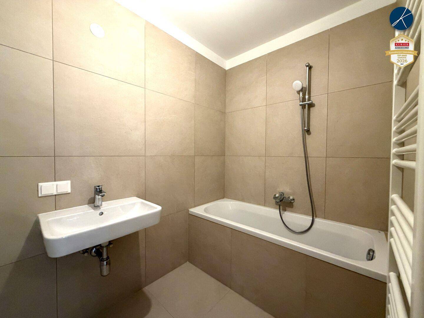 Modernes Badezimmer mit Badewanne, Waschbecken und beigefarbenen Fliesen, ausgestattet mit Handtuchheizkörper.