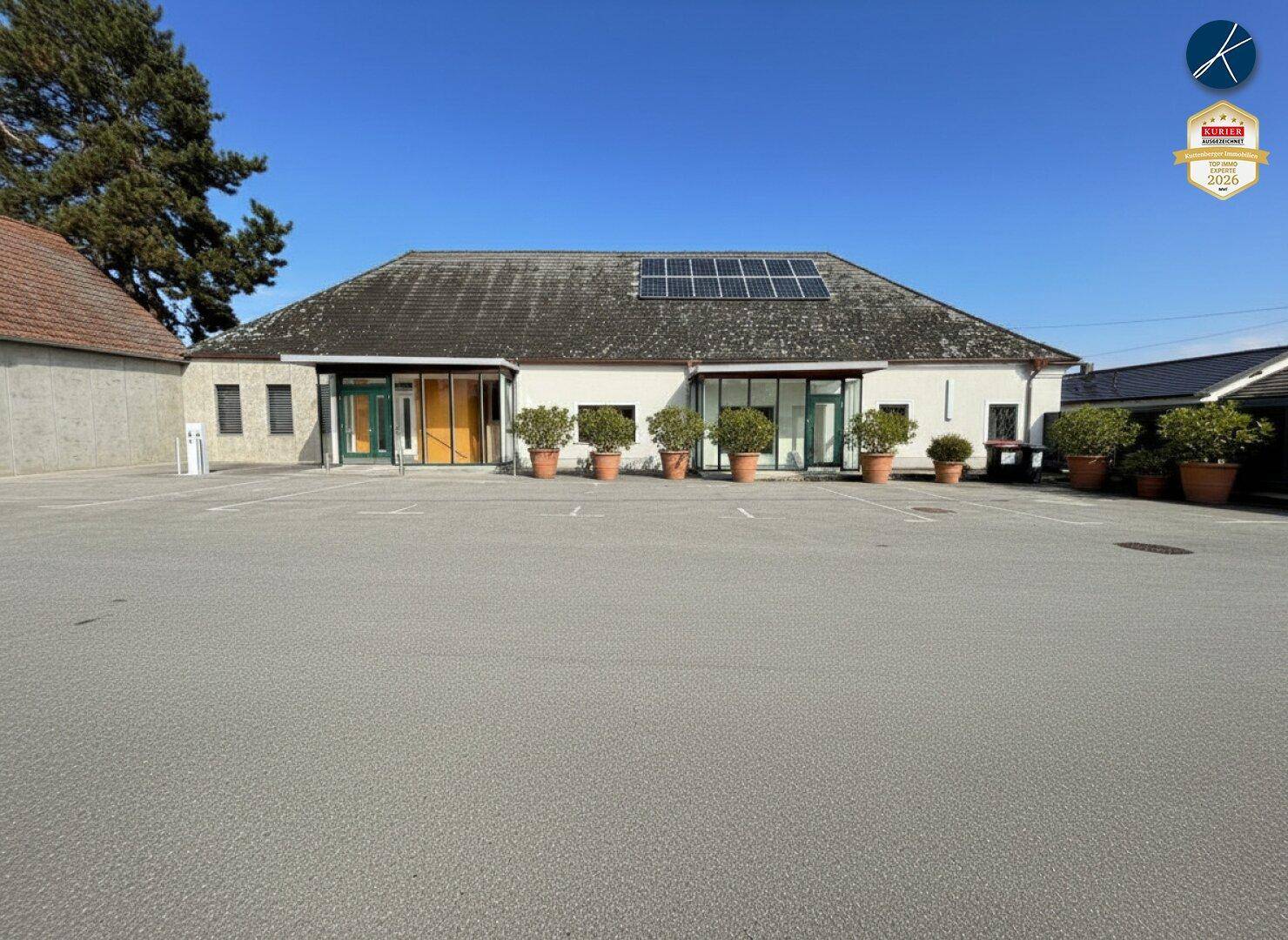 Großes Gebäude mit weitläufigem Parkplatz, Solaranlage auf dem Dach und gepflegten Pflanzenkübeln.