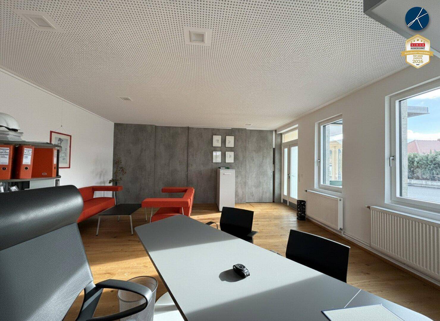 Modern eingerichtetes Büro mit Holzboden, großen Fenstern und orangefarbenen Sitzmöbeln.