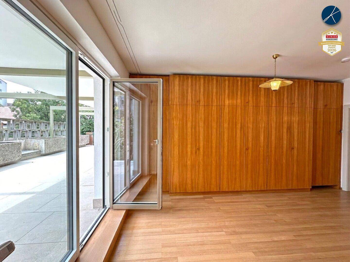 Geräumiger Wohnbereich mit Holzboden, großen Fenstern und integrierten Holzschränken mit Terrassenzugang.