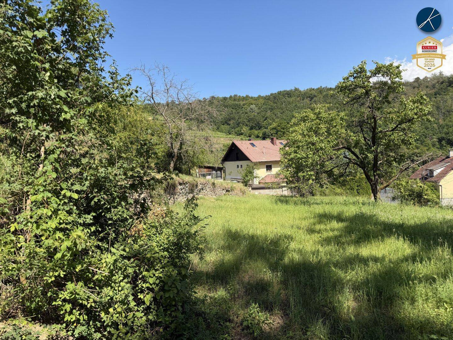 Haus in ländlicher Umgebung, umgeben von grünen Wiesen und Bäumen unter blauem Himmel.