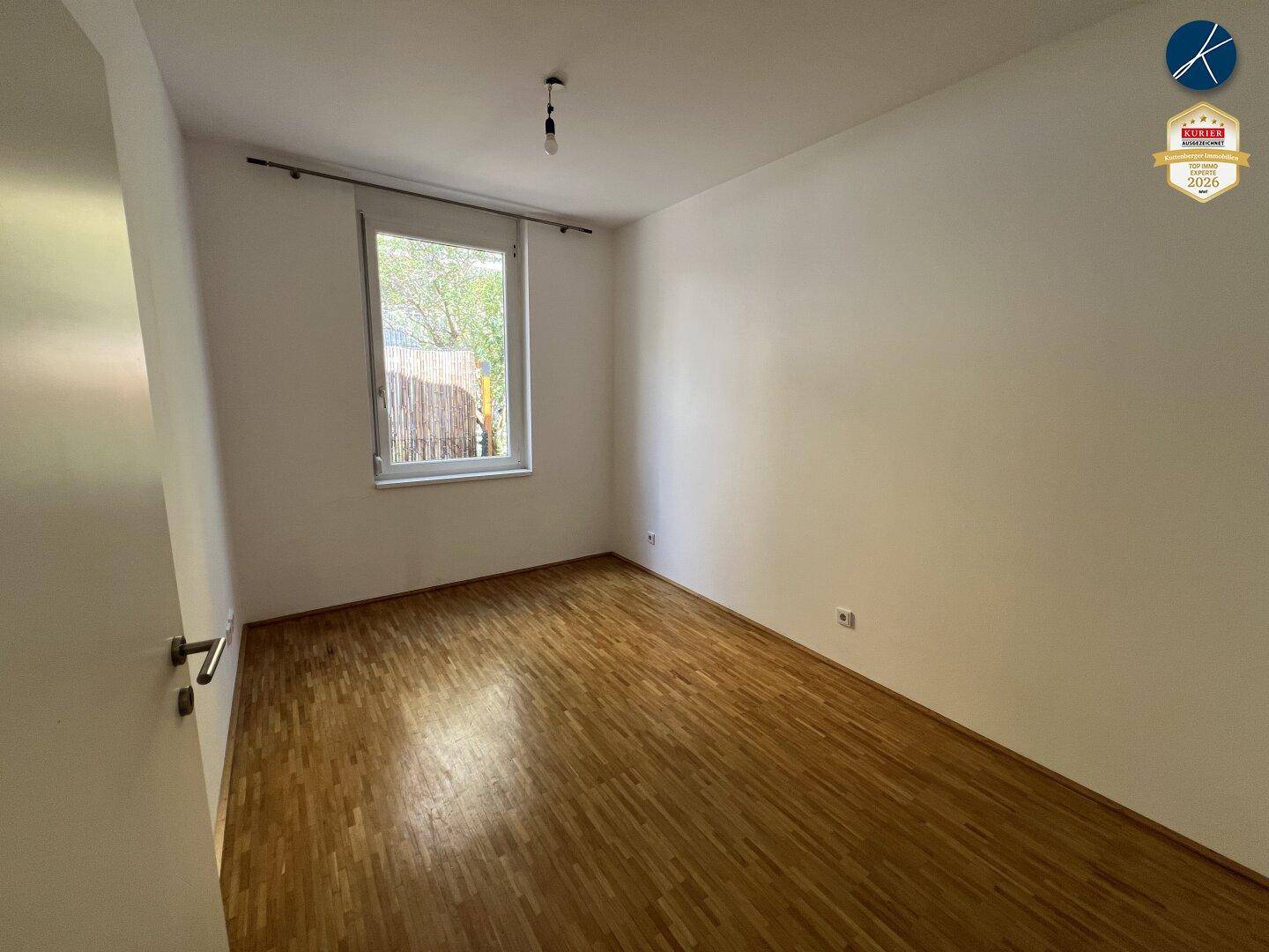 Helles, leeres Zimmer mit Holzparkettboden und einem Fenster mit Blick ins Grüne.