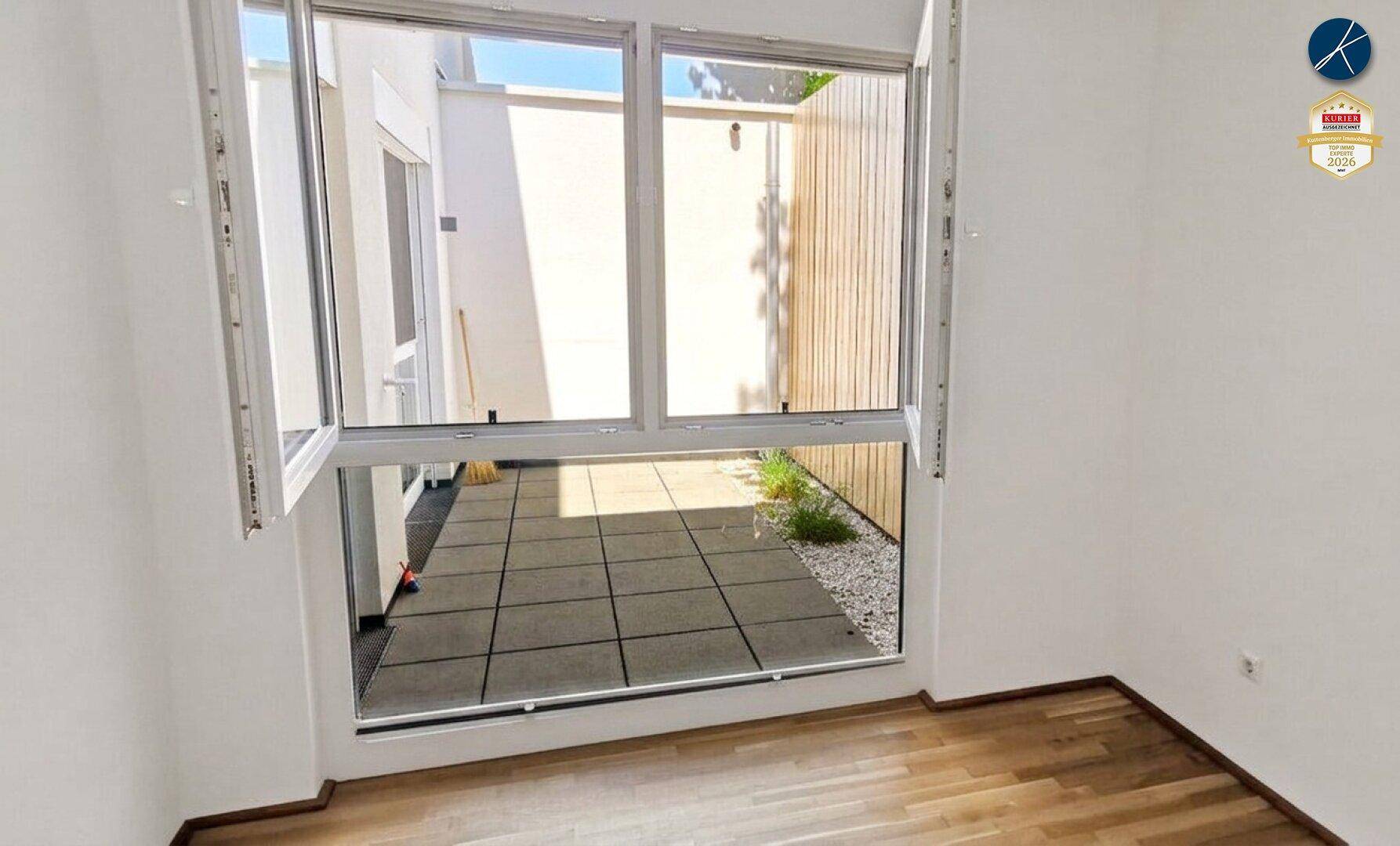 Heller Innenraum mit Holzboden und großem Fenster, das auf eine kleine gepflasterte Terrasse führt.