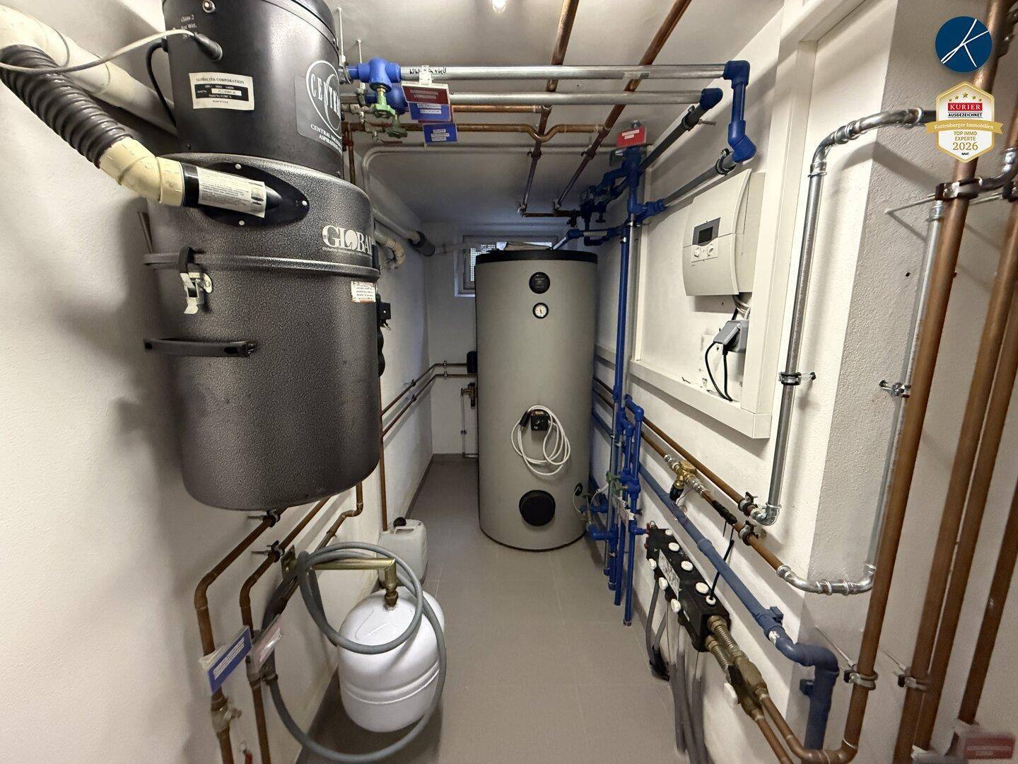 Technikraum mit Heizungsanlage, Warmwasserspeicher und sichtbaren Rohren an der Decke.