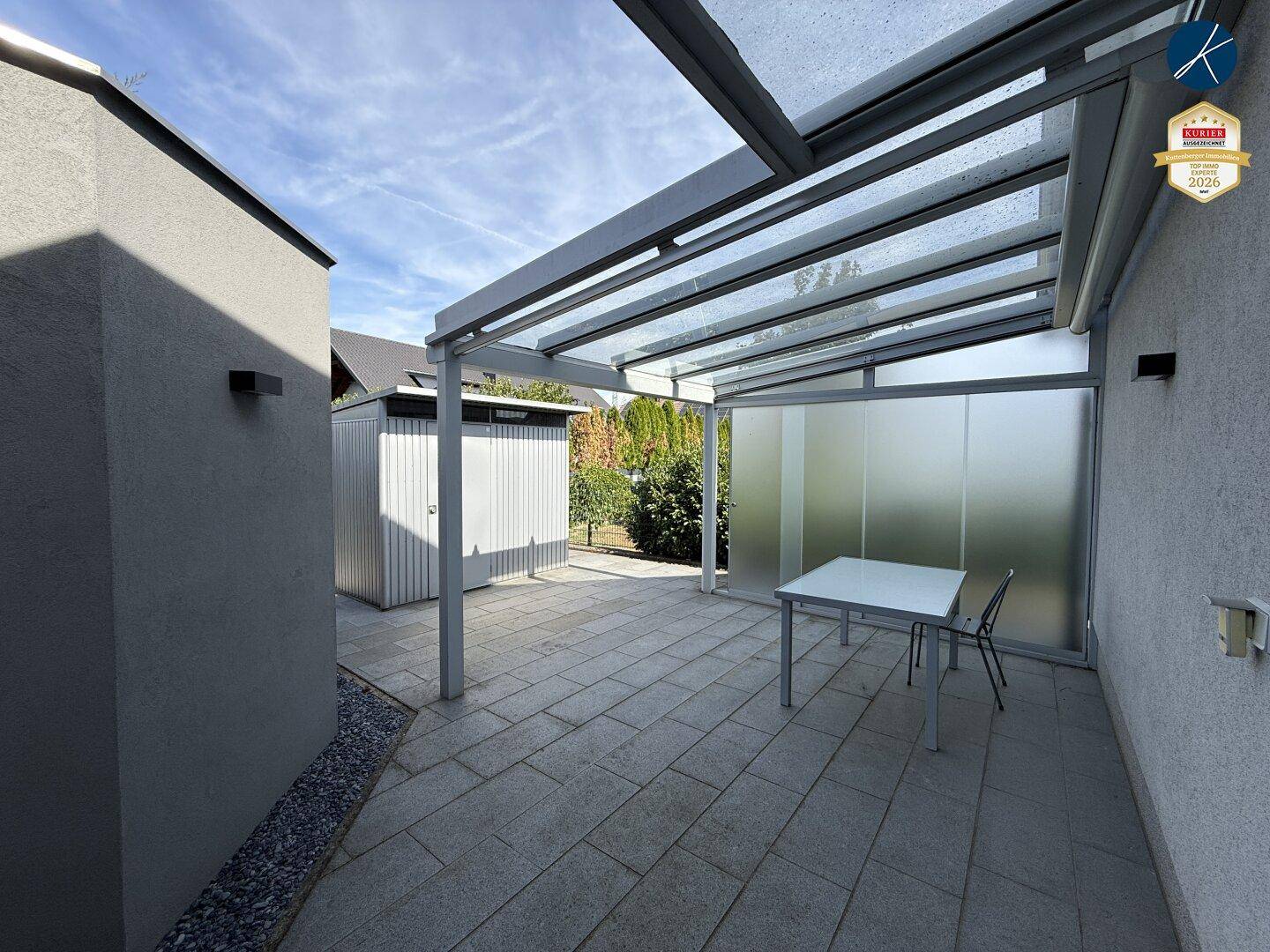 Überdachte Terrasse mit Sitzgelegenheiten, Glasdach und Blick in den Garten.