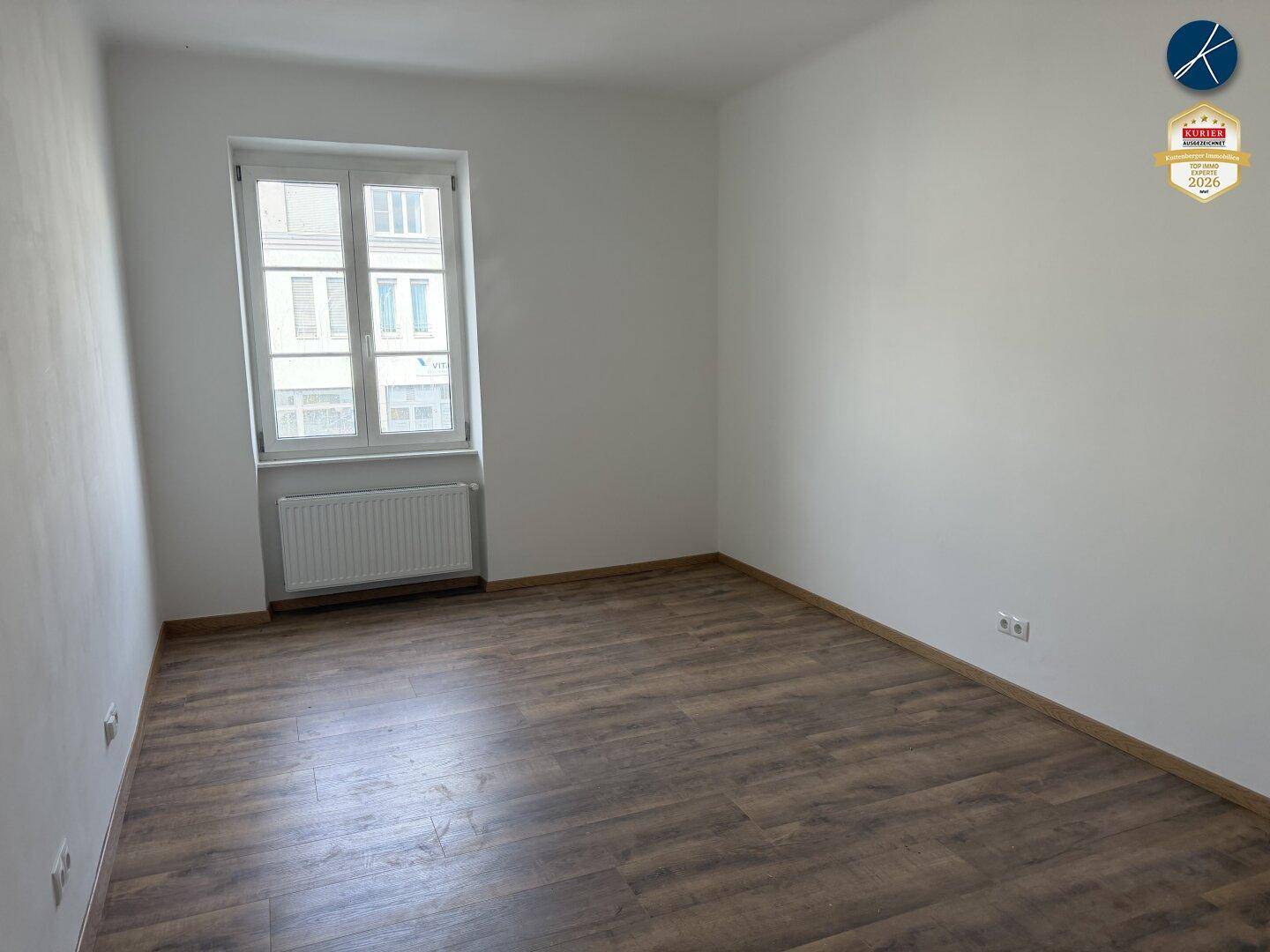 Leerer, heller Raum mit Laminatboden und einem großen Fenster, das Tageslicht spendet.