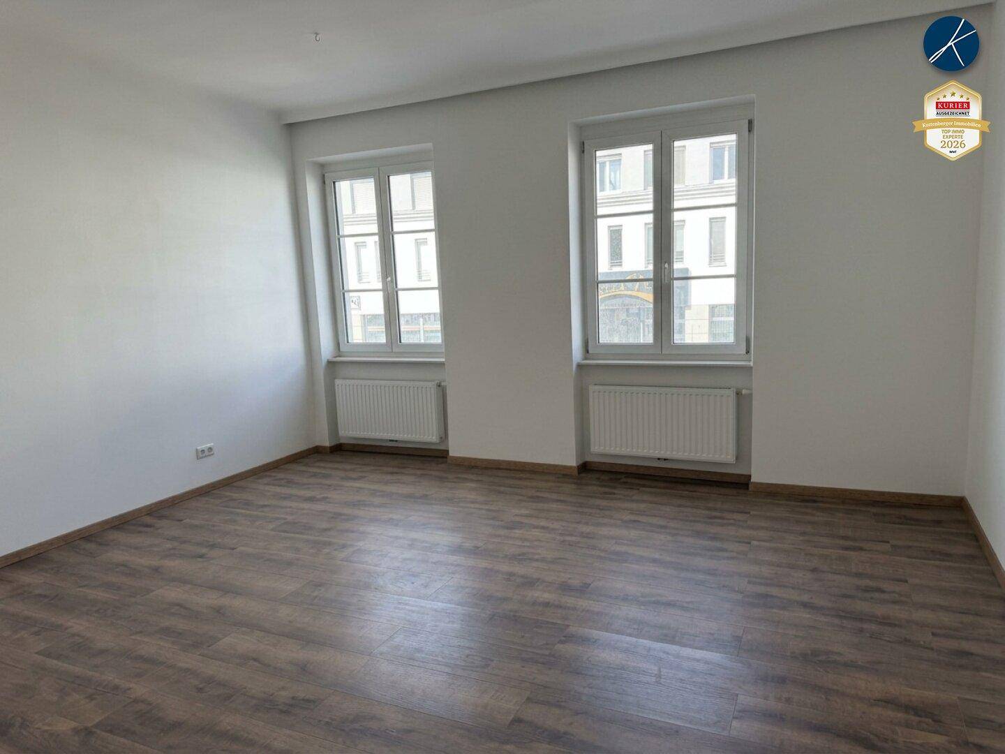 Heller, leerer Raum mit Laminatboden und zwei großen Fenstern, die viel Tageslicht hereinlassen.