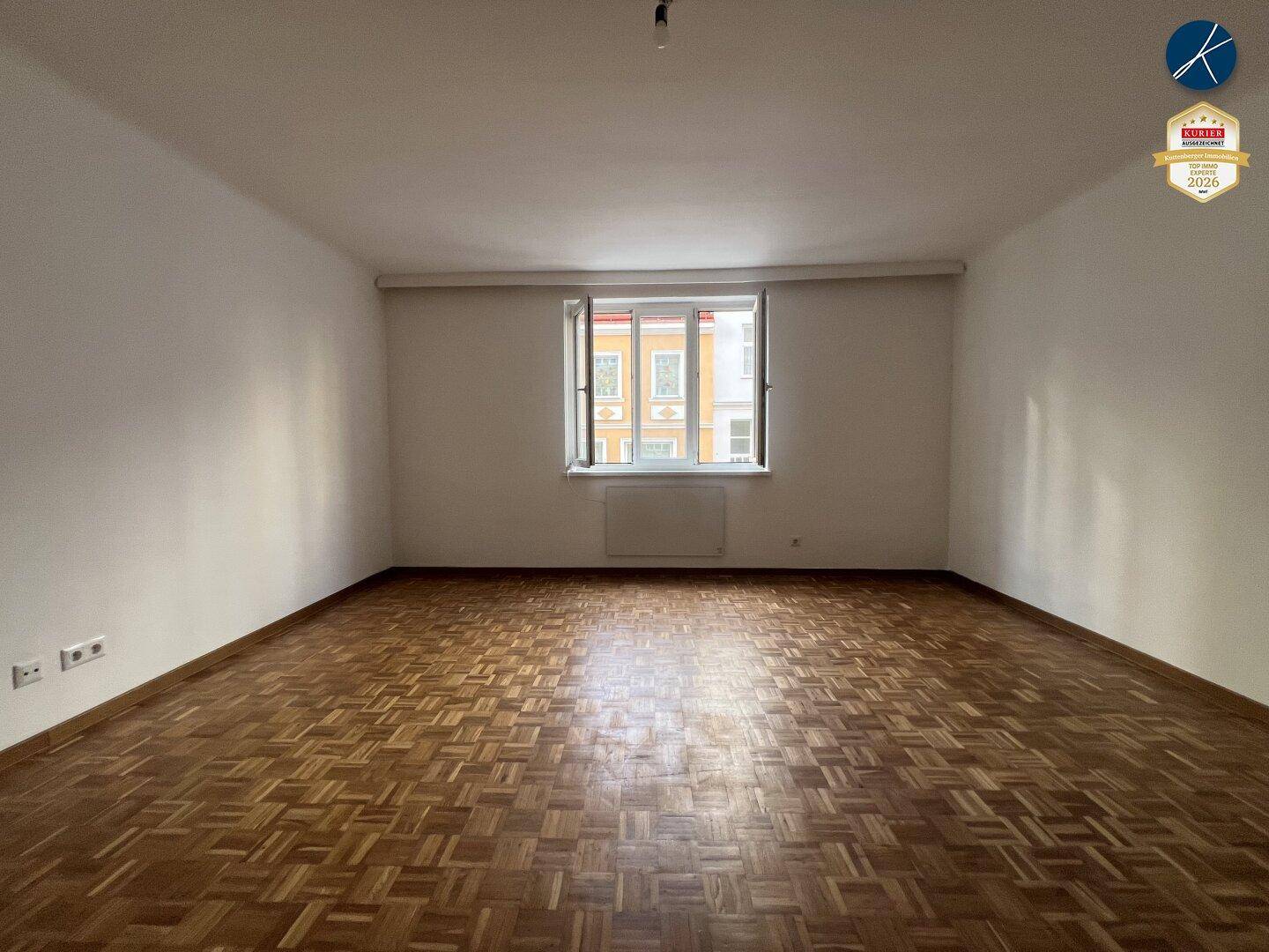 Geräumiges, leeres Zimmer mit Fischgrätparkett und einem Fenster, das auf die Stadt blickt.