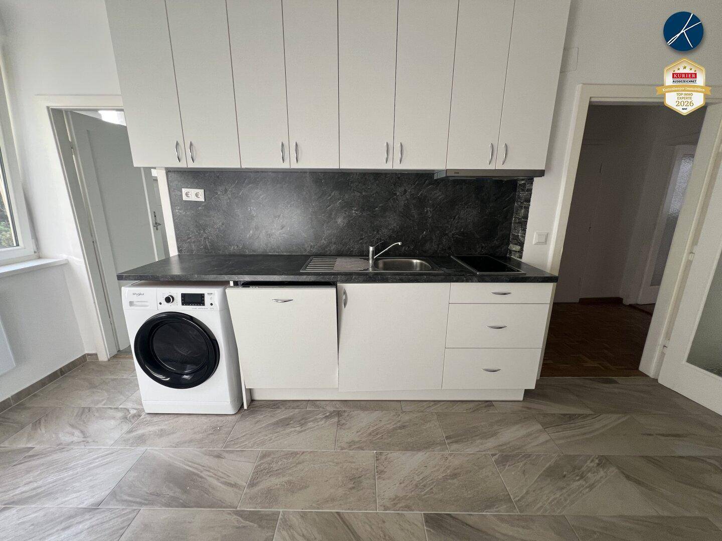 Moderne Küche mit weißen Oberschränken, dunkler Arbeitsplatte, Spüle und integrierter Waschmaschine.