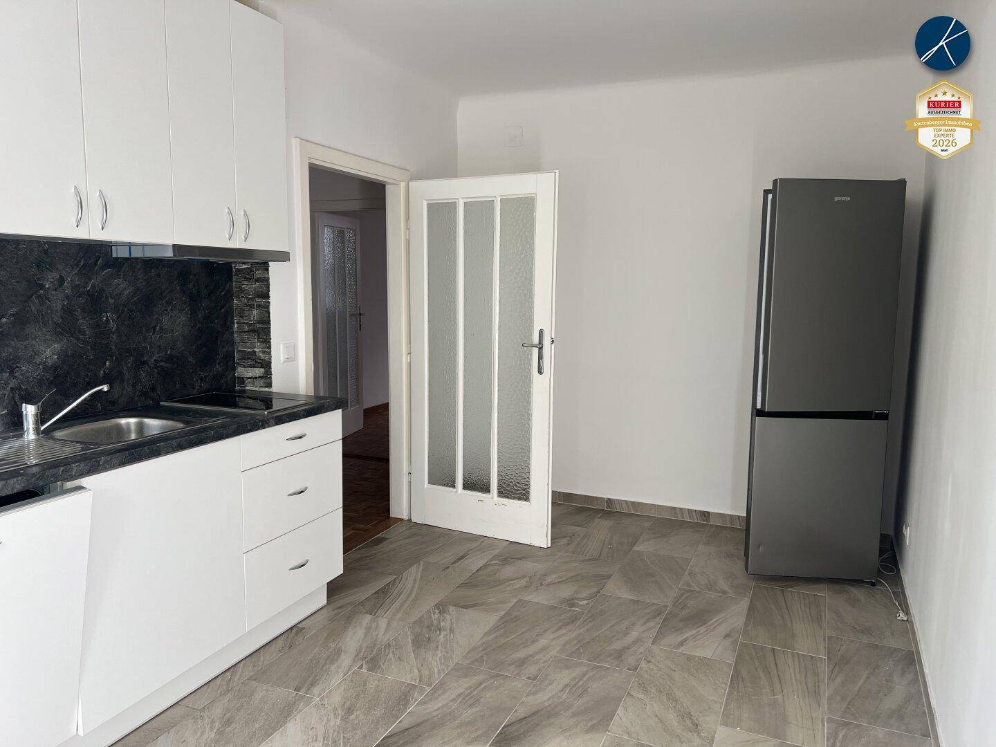 Funktionale Küche mit weißen Schränken, dunkler Arbeitsplatte, Kühlschrank und Zugang zu einem weiteren Raum.