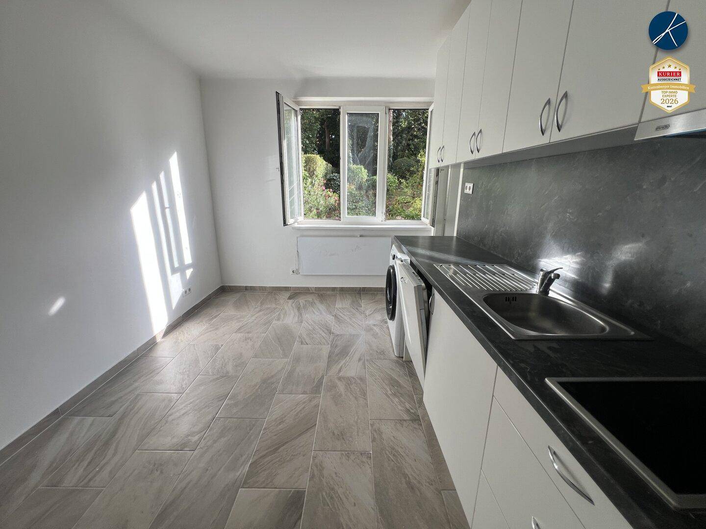 Helle Küche mit Fliesenboden, weißen Schränken und Sonnenlicht, das durch das Fenster fällt.