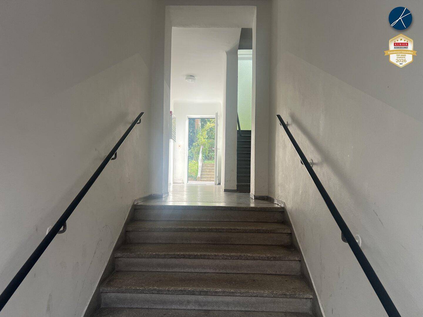 Treppenhaus mit robusten Betonstufen und schwarzen Handläufen, das zu einem hellen Eingangsbereich führt.