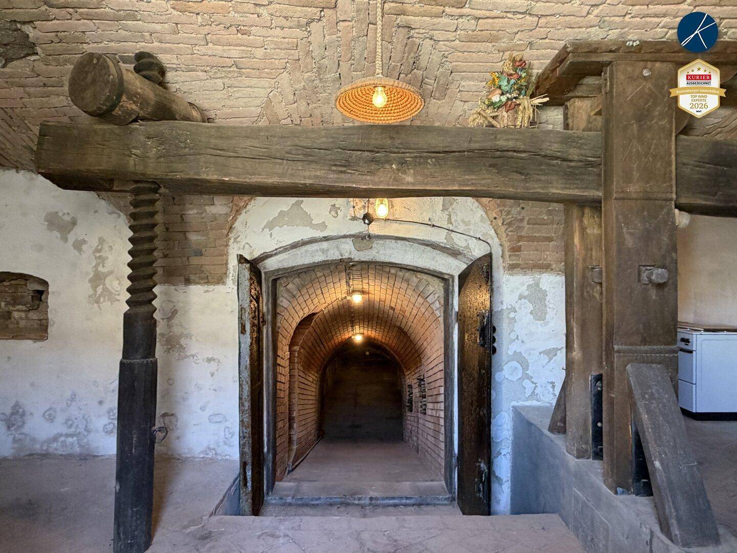 Historischer Weinkeller mit großer Holzpresse und gewölbtem Eingang zu weiteren Gängen.