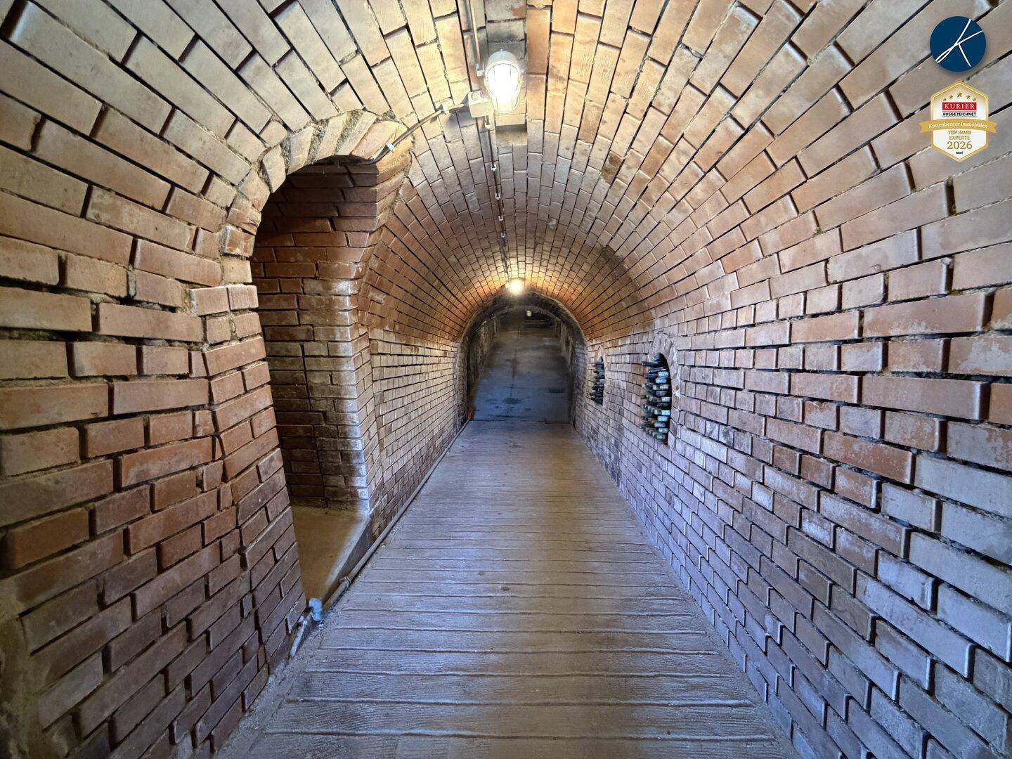Langer, gewölbter Backsteintunnel im Keller mit Holzboden und Beleuchtung.