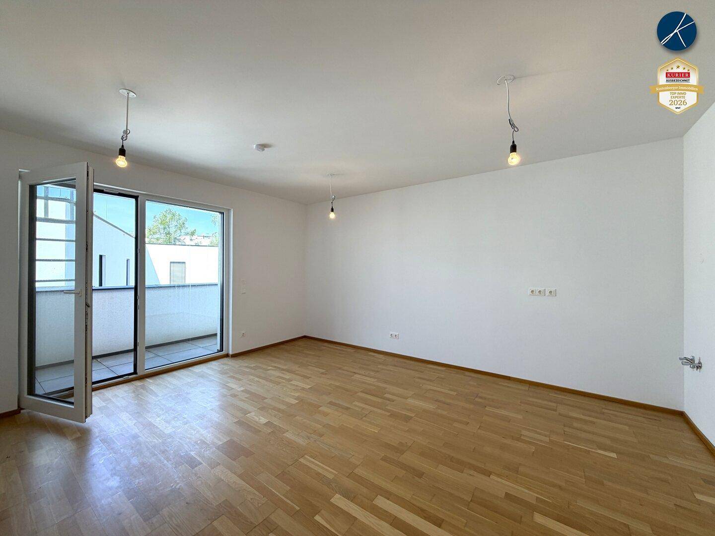 Geräumiger, leerer Wohnbereich mit hellem Parkettboden und großem Fenster zum Balkon.
