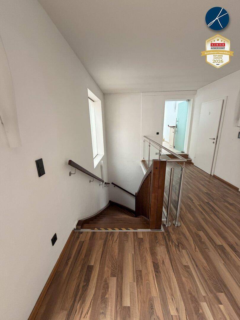 Treppenhaus mit Holzstufen und modernem Glas-Metall-Geländer, das zu einer unteren Etage führt.