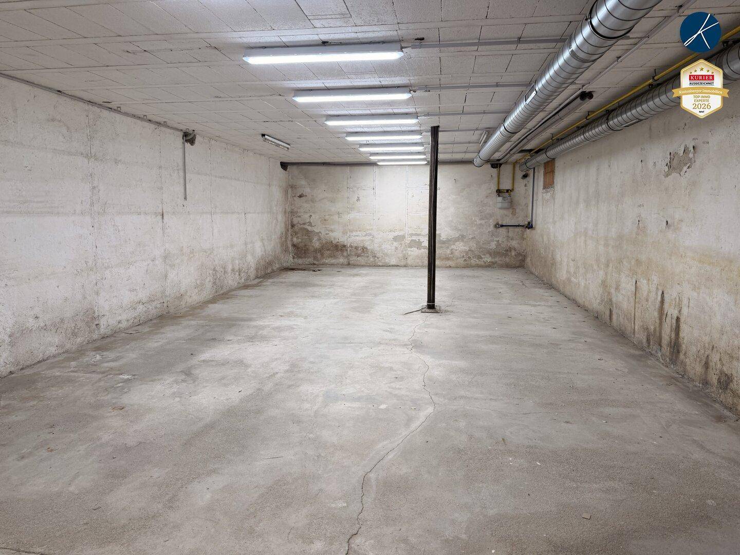 Geräumiger Kellerraum mit Betonboden und -wänden, sichtbaren Rohren an der Decke und Leuchtstoffröhren.