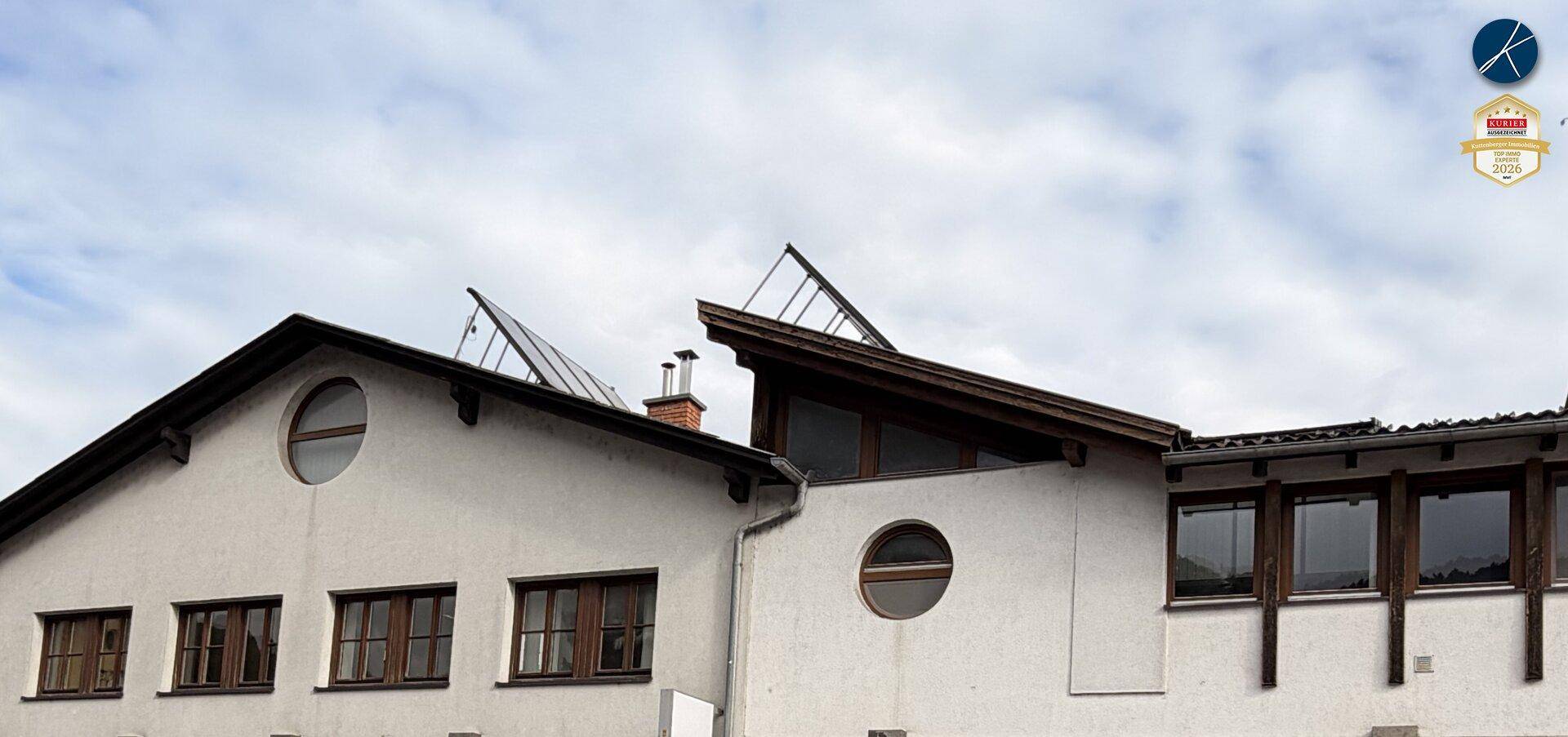 Dachlandschaft eines Gebäudes mit runden und rechteckigen Fenstern sowie sichtbaren Solarpaneelen auf dem Dach.