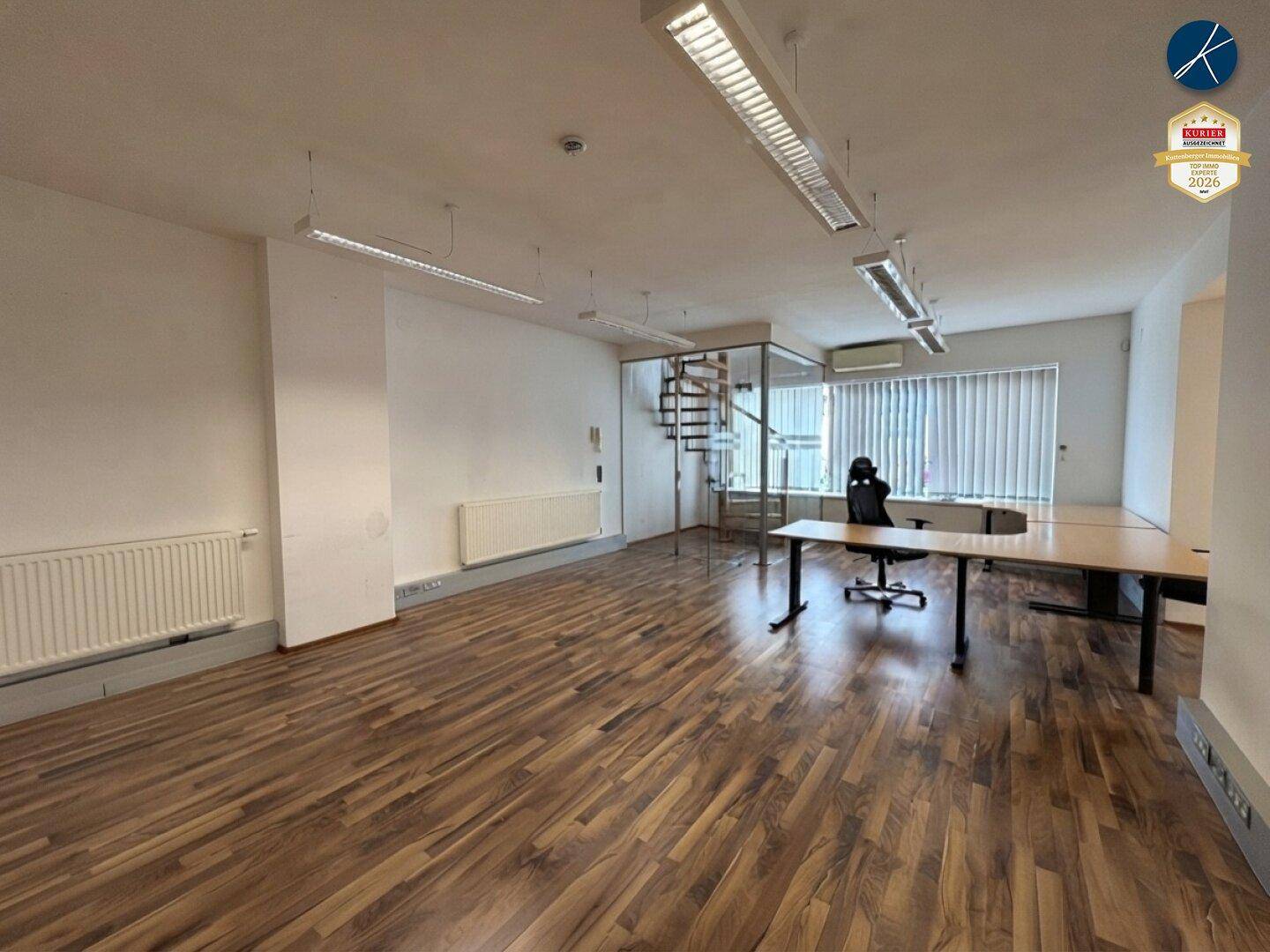 Großer, offener Bürobereich mit Laminatboden, weißen Wänden, Deckenleuchten und einer Wendeltreppe im Hintergrund.