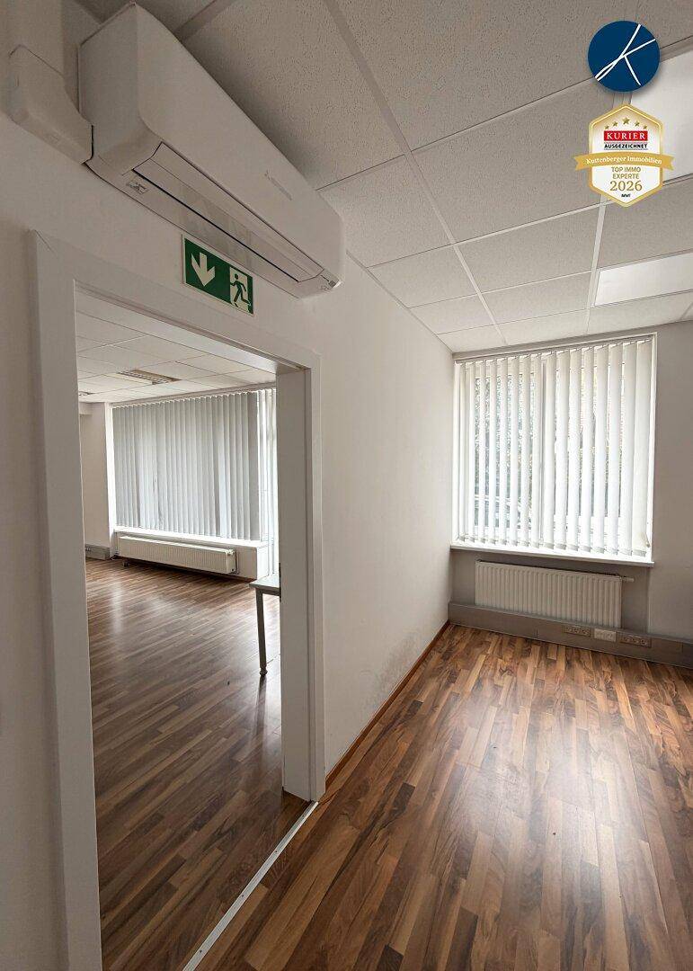 Heller Büroraum mit Laminatboden, weißen Wänden, Fenster mit Jalousien und einer Klimaanlage an der Decke.