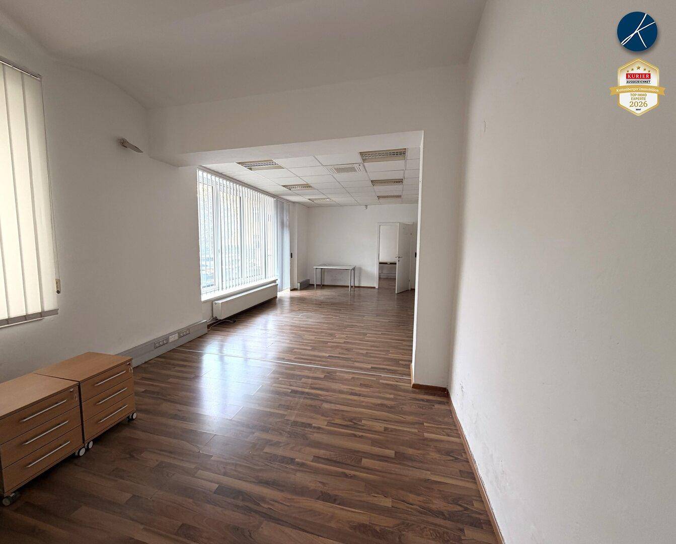 Leerer Büroraum mit Laminatboden, weißen Wänden und großen Fenstern mit Jalousien, bereit zur Einrichtung.
