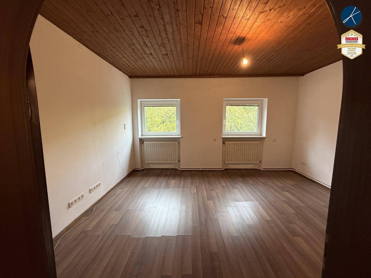 Leerer Raum mit dunklem Laminatboden, Holzdecke und zwei Fenstern mit Heizkörpern darunter.