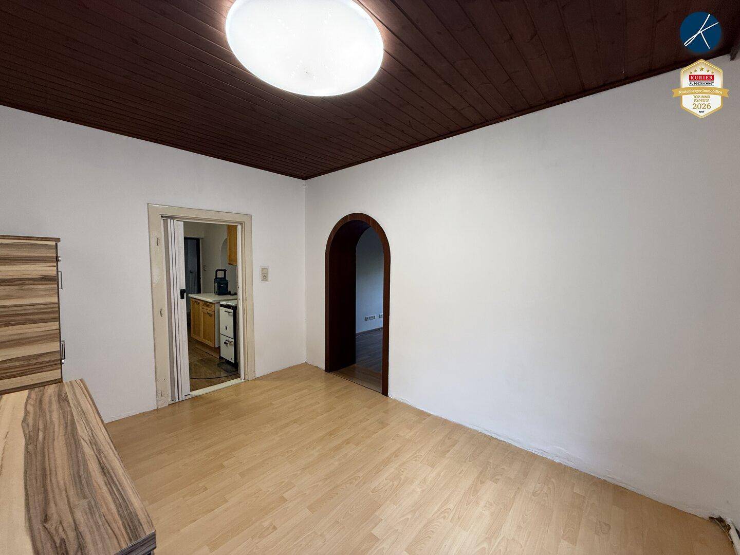 Leerer Raum mit hellem Laminatboden, weißen Wänden und dunkler Holzdecke, mit Zugang zu weiteren Zimmern.