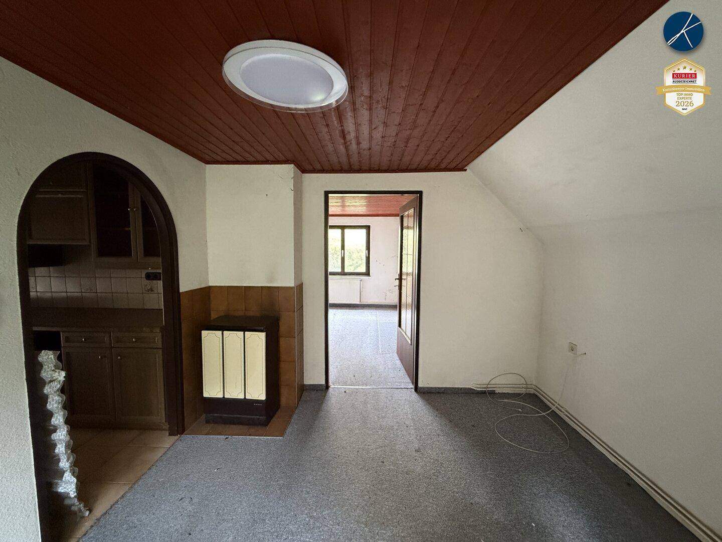 Wohnraum mit grauem Teppichboden, Holzdecke, Kachelofen und Zugang zu einer kleinen Kochnische.