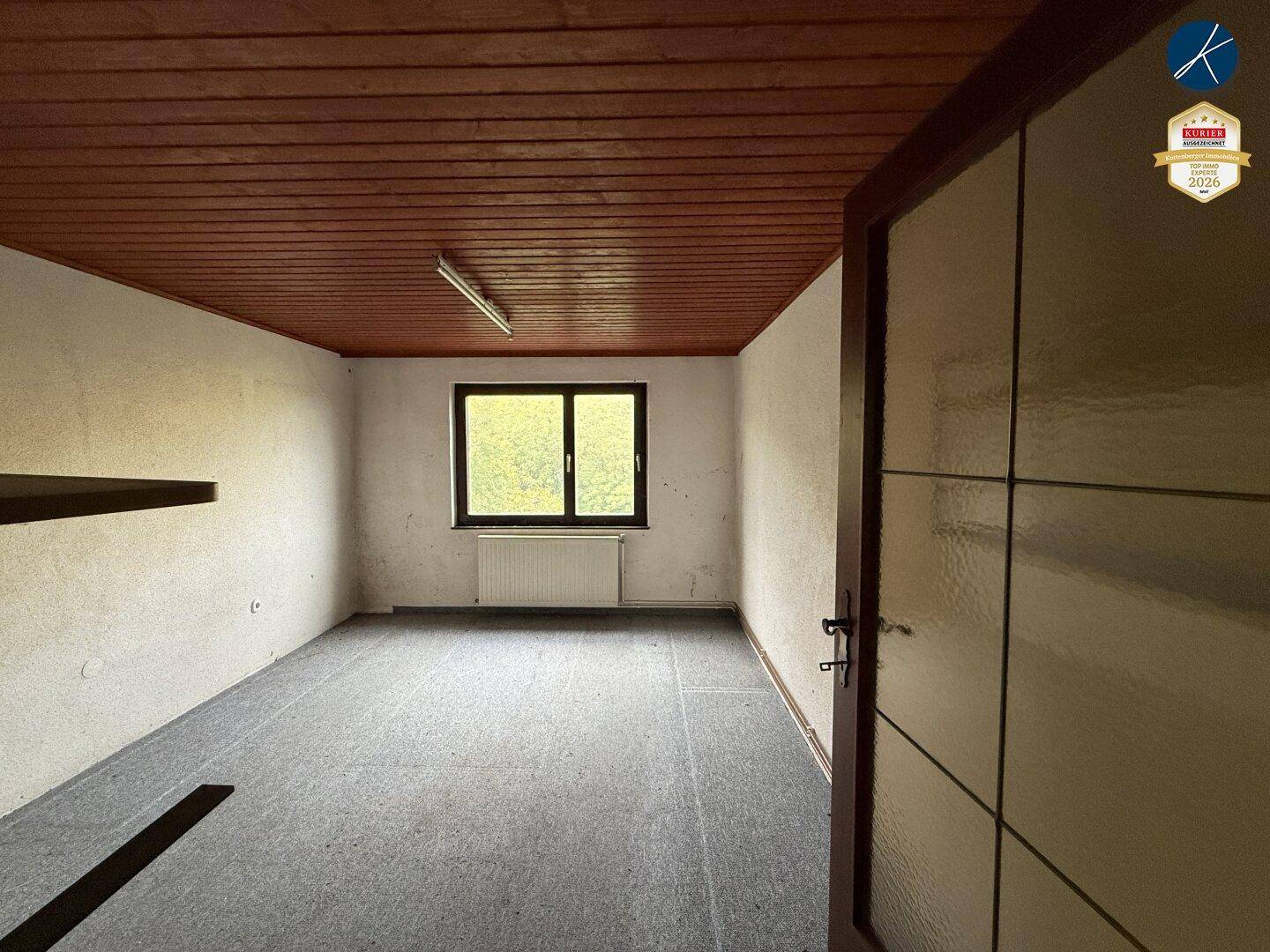 Leerer Raum mit grauem Teppichboden, Holzdecke und einem Fenster mit Blick ins Grüne.